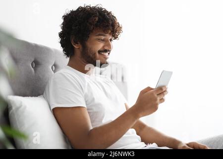 Positive indian guy scrolling in bed, using smartphone Stock Photo