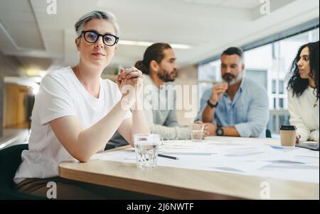Mature businesswoman looking at the camera while sitting in a meeting with her team. Group of creative design professionals discussing a set of bluepr Stock Photo