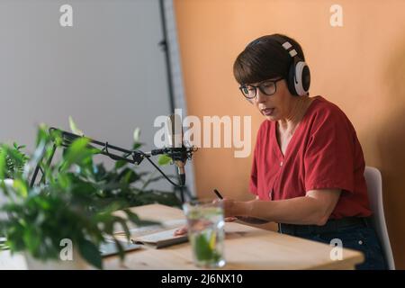 Mature woman making podcast recording for her online show. Attractive business woman using headphones front of microphone for a radio broadcast Stock Photo