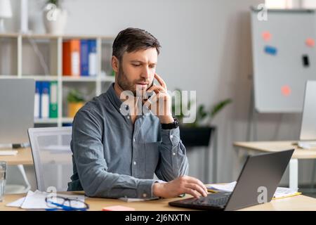 Focused mature businessman having phone conversation with client, sitting at office, typing on laptop keyboard Stock Photo