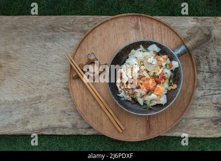 Stir-fried wide rice noodles with Bacon, Egg, Dried shrimp in small steaming iron pot and wooden chopsticks served on round wooden tray. Selective foc Stock Photo