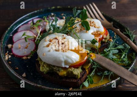 Bread toast, poached eggs, avocado pulp, pea microgreens and fresh vegetables on plate in cafe, close up, breakfast time Stock Photo