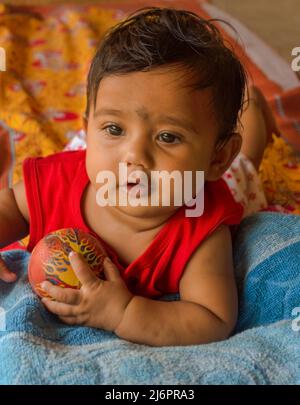 Cute Indian baby playing with a ball lying on a yellow printed bed sheet over a mat. Selective focus on baby's face Stock Photo