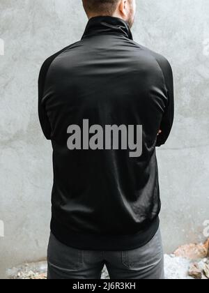 Young man with folded arms in a black sports jacket. Rear view on a background of light gray concrete wall. Design and layout of men's uniforms for printing. Stock Photo