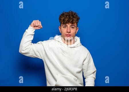Curly haired young man in his 20s wearing white sweatshirt sticking out biceps demonstrating strength power isolated on pastel blue color background Stock Photo