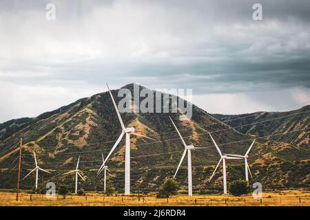 Wind turbins in a field with mountains in the background under overcasst skies. Powerlines are also strung around - Idaho USA Stock Photo