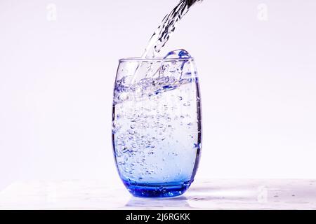 Pouring purified drinking water into a blue glass, white background carrara marble base. full glass. Stock Photo
