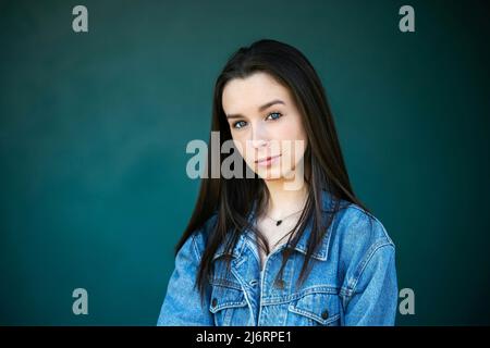 A beautiful teen brunette girl with a serious look leaning against a wall in thought with a teal green turquoise background Stock Photo