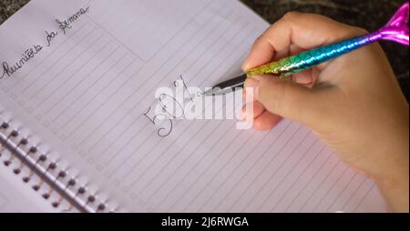50 percent handwritten, female hand holding a colored pen in the shape of a mermaid's tail, in a white spiral notebook, on a granite table Stock Photo