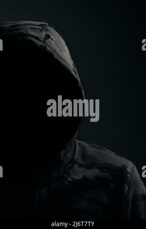 Man in dark camouflage hoodie with covered face dark background, hacker bandit and attacker Stock Photo