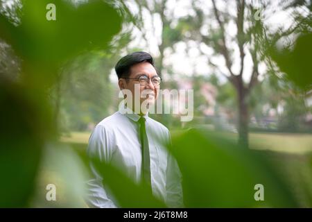 Well dressed mature Asian businessman smiling. Outdoor nature with residential property background. Stock Photo