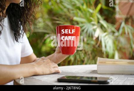 Woman holding a red cup with text Self care, and a book on table. Outdoor with palm tree background. Stock Photo