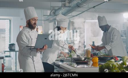 Man and woman using laptop to cook food recipe in kitchen, making gourmet dish. Team of people cooking professional authentic meal with computer online website, preparing ingredients. Stock Photo