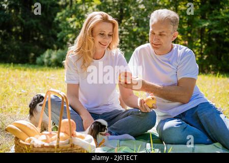 Mature couple lie with their dog in park. An elderly couple is resting in nature with dog. Close-up portrait of an elderly man and woman in white shirts and jeans. Stylish and modern grandparents. Stock Photo