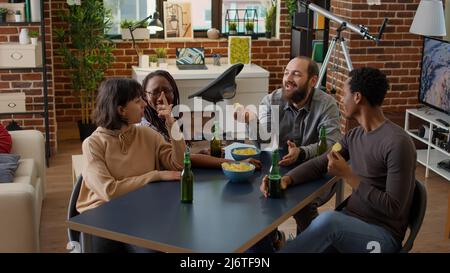 Diverse group of friends talking and having fun in living room, drinking beer and eating chips. Happy people having conversation at leisure activity gathering, enjoying discussion together. Stock Photo