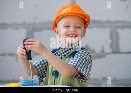 Kid boy twists bolt with screwdriver. Child repairman with repair tool. Child in helmet and boilersuit on construction site. Little worker engineer. Stock Photo