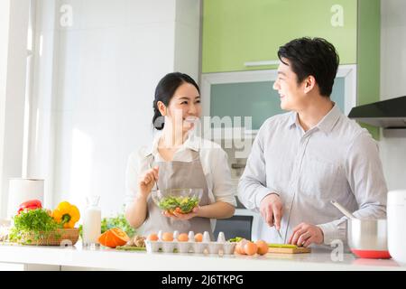 In the family kitchen, a young couple cooks together, the husband chopping vegetables and the wife stirring salad greens - stock photo Stock Photo