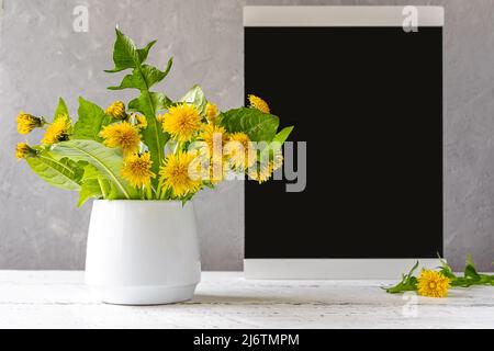 Mock up creation. Black chalk desk in frame on white wooden table with grey concrete background and dandelions in vase Stock Photo