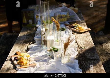 Wedding party banquet outdoors in forest. Dining table decorated in boho style with candles, white cloth, flowers, served with plates, tableware Stock Photo