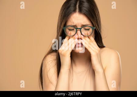 Sinus pain, sinus pressure, sinusitis. Sad woman with glasses, holding her nose because sinus pain on a beige background. Stock Photo