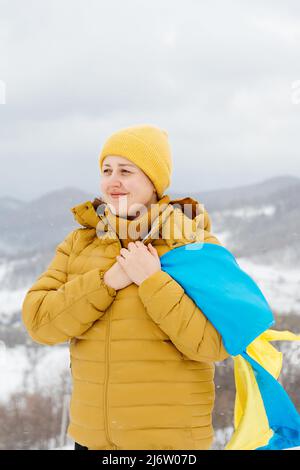 Young girl holding a blue and yellow national flag of Ukraine on a background of snowy Carpathian mountains and blue sky. Woman in orange jacket and yellow hat. Constitution day. Stock Photo