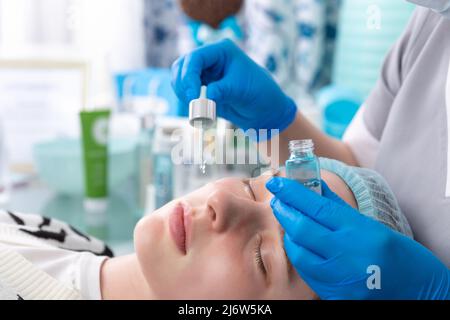 Hands of cosmetologist dripping nourishing oil facial skin of young woman in spa salon. Facial treatment and skincare concept. Stock Photo