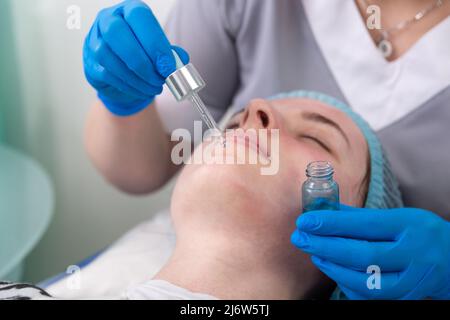 Hands of cosmetologist dripping nourishing oil facial skin of young woman in spa salon. Facial treatment and skincare concept. Stock Photo