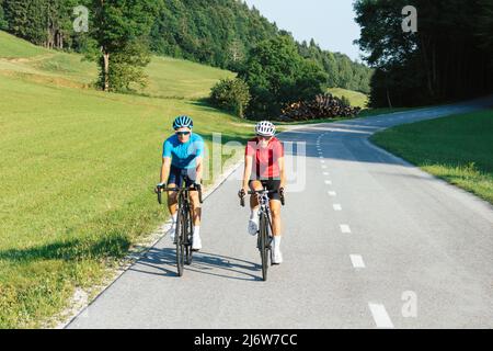 Front view of a woman and man, professional racing bikers in sportswear, riding along an asphalt road in the beautiful green nature. Stock Photo
