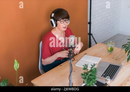 Mature woman making podcast recording for her online show. Attractive business woman using headphones front of microphone for a radio broadcast Stock Photo