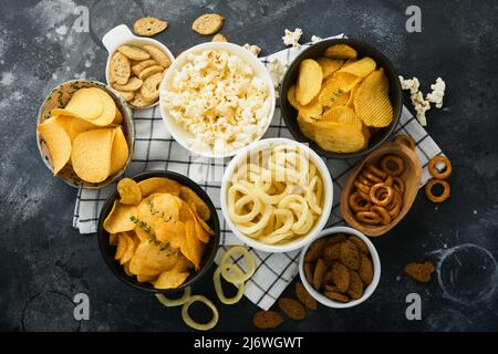 Unhealthy food. Snacks. All classic potato snacks with peanuts, popcorn and onion rings and salted pretzels in bowl plates on black concrete backgroun Stock Photo