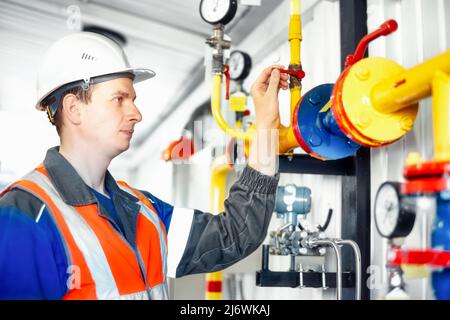 Engineer in white helmet and overalls shuts off natural gas supply tap through pipes in gas boiler room. Worker maintains boiler room equipment. Real scene Stock Photo