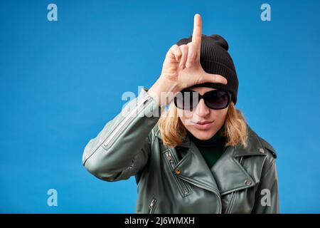 Portrait of young woman with blonde hair in green jacket standing, showing loser gesture with hand on forehead. Stock Photo