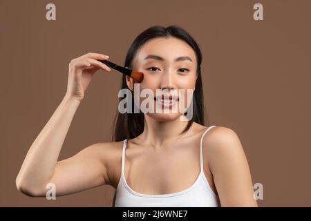 Attractive young asian lady with makeup brush applying blush on her cheeks, smiling at camera over brown background Stock Photo