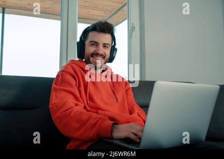 Young man wearing red hoodie sitting on couch in living room at home enjoying studying using laptop and headset looking at the camera with big smiles. Stock Photo