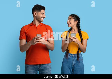 Cheerful Young Arabic Couple Holding Smartphones And Looking At Each Other Stock Photo