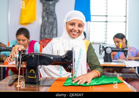 Happy smiling muslim woman in hijab loooking at camera while stitching using sawing machine at garments - concept of equality, diversity and Stock Photo