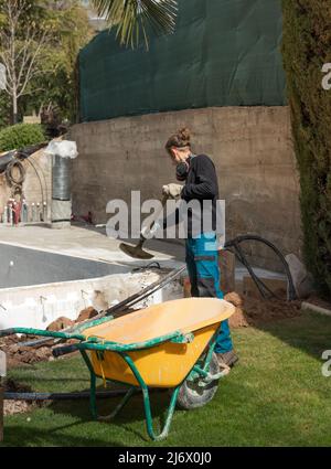 Young labourer working outdoors on a pool site in a single-family house along with a yellow wheelbarrow and shovel Stock Photo