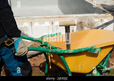 Unrecognizable man construction worker holding a yellow wheelbarrow and a shovel surrounded by construction materials Stock Photo