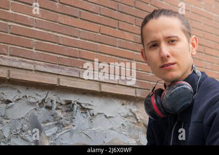 Portrait of tired labourer man working in construction looking at the camera with headphones on and braick behind Stock Photo