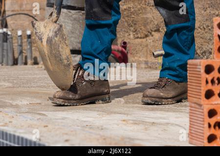 Worker's legs working in construction site with a shovel resting on safety boots and bricks around Stock Photo