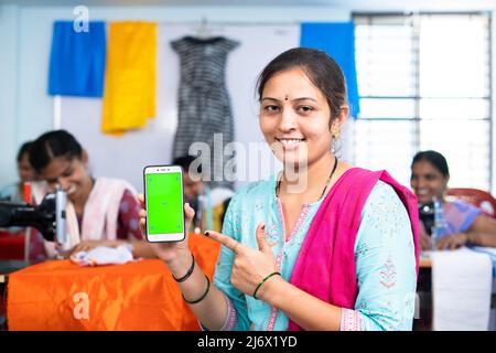 Smiling garment owner showing green screen mobile phone by pointing finger by looking camera - concept of online booking, advertisement and small Stock Photo