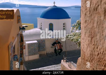 Oia, Santorini, Greek Islands, Greece - Man playing accordion next to pretty Greek Orthodox church with blue dome in dazzling bright sunslight Stock Photo