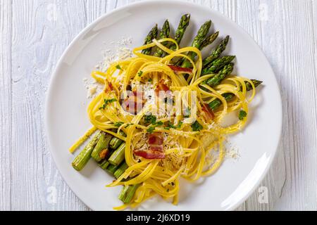 roasted asparagus spears with spaghetti sprinkled with shredded cheese, fried bacon pieces and parsley on white plate on white textured wood table, fl Stock Photo