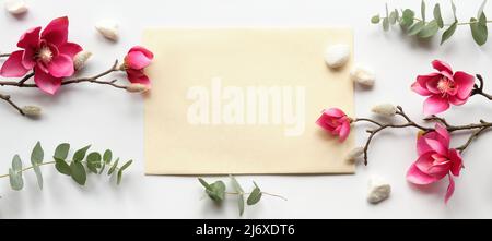 Pink magnolia flowers on twigs, fresh eucalyptus leaves. Blank pink card with grey envelope. Stock Photo