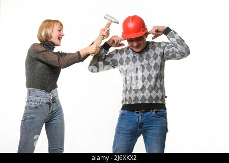 The wife screams and hits her husband on the head with a hammer, the husband is wearing a helmet and he covered his ears with his fingers Stock Photo