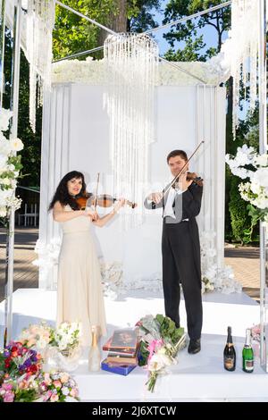 Man and woman playing violins under the wedding arch. Stock Photo