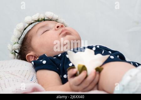 close-up detail of the face of a beautiful latin baby girl resting on a white pillow. holding in her right hand a white flower symbol of her purity an Stock Photo