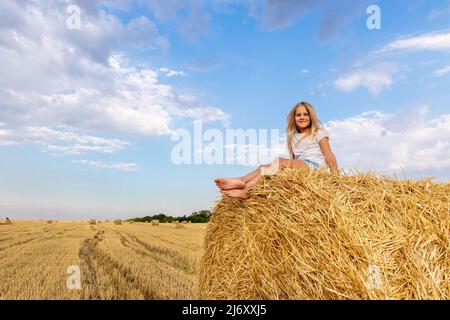 Portrait of cute little blond beautiful adorable cheerful caucasian kid girl enjoy sitting on hay stack or bale on harvested wheat field warm summer Stock Photo