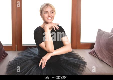 a young, smiling ballerina in a black tutu sits in front of the window with her chin tucked in Stock Photo