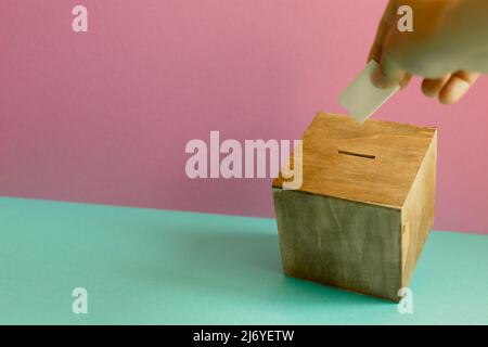Hand putting voting paper in Ballot box on mint green table. pink wall background Stock Photo